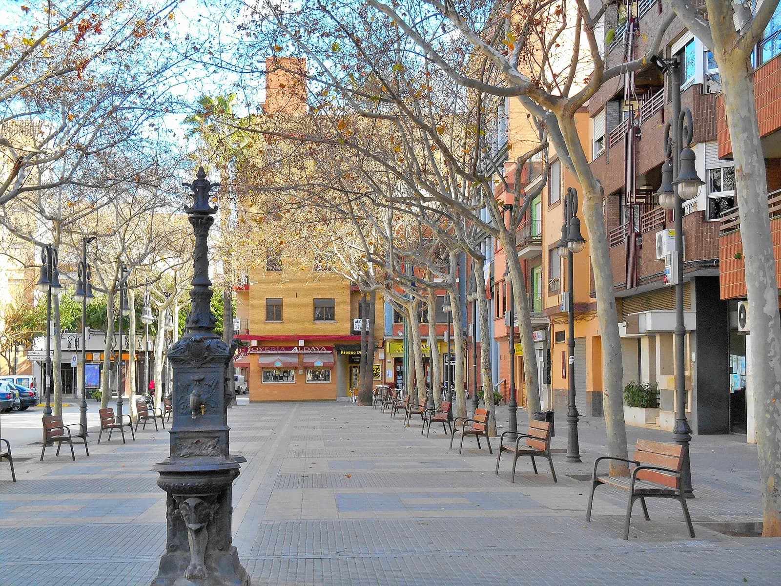 Plaça de la Constitució de Aldaia, paseo peatonal con plátanos, bancos y una fuente de hierro, con edificios de viviendas a los lados