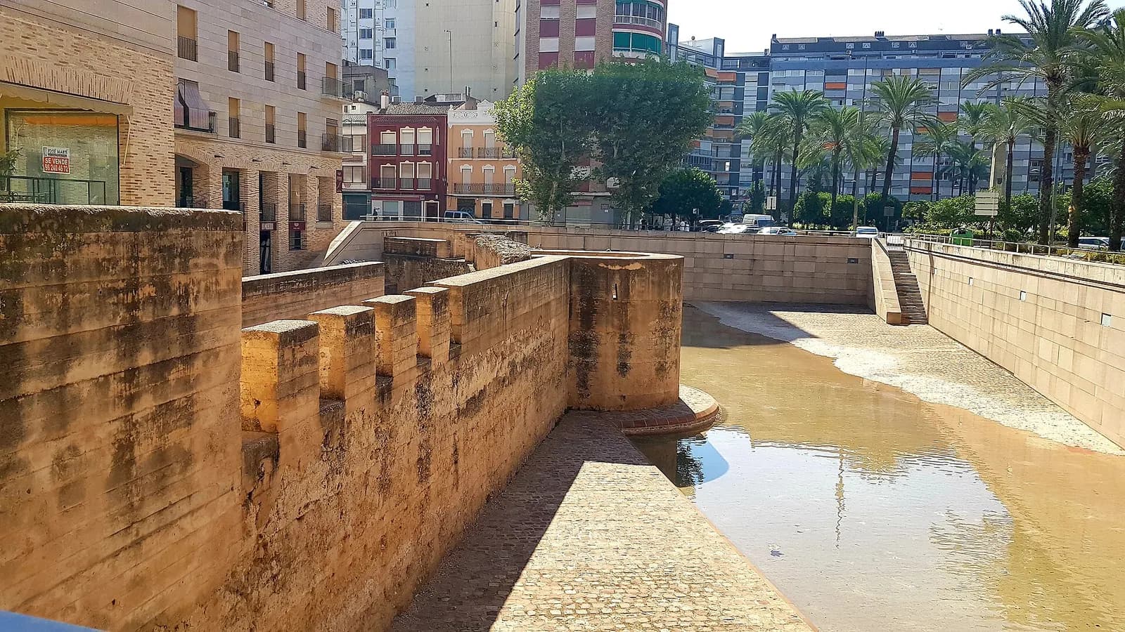 Muralla árabe de Alzira junto a un antiguo cauce, con edificios de viviendas y palmeras detrás