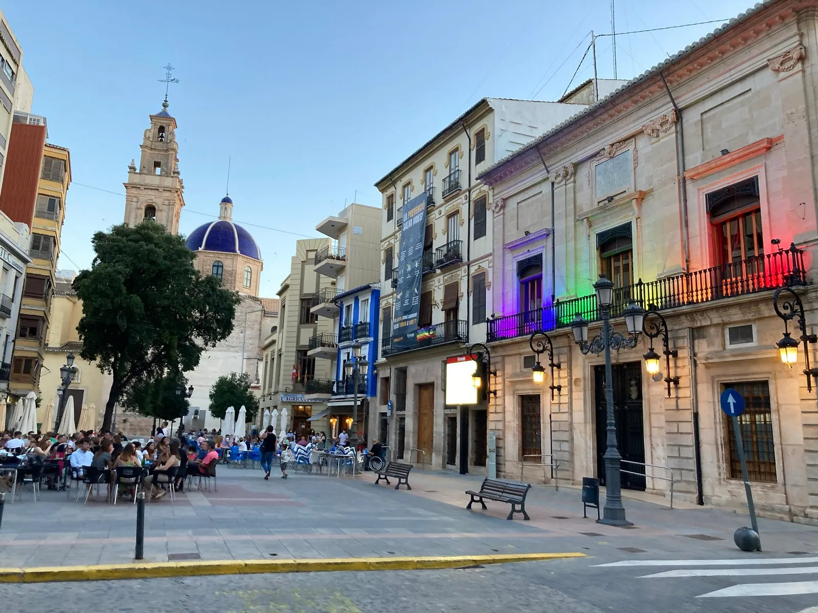 Plaça de l'Ajuntament de Sueca con terrazas, edificios históricos y el campanario y la cúpula azul de la iglesia al fondo