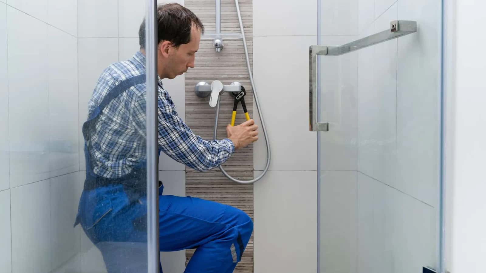 Fontanero con peto azul ajustando con unos alicates el grifo monomando de una ducha con mampara